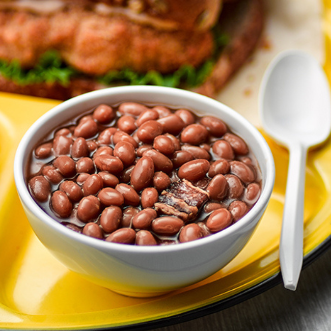 A bowl of baked beans on a yellow plate next to a plastic spoon and a sandwhich