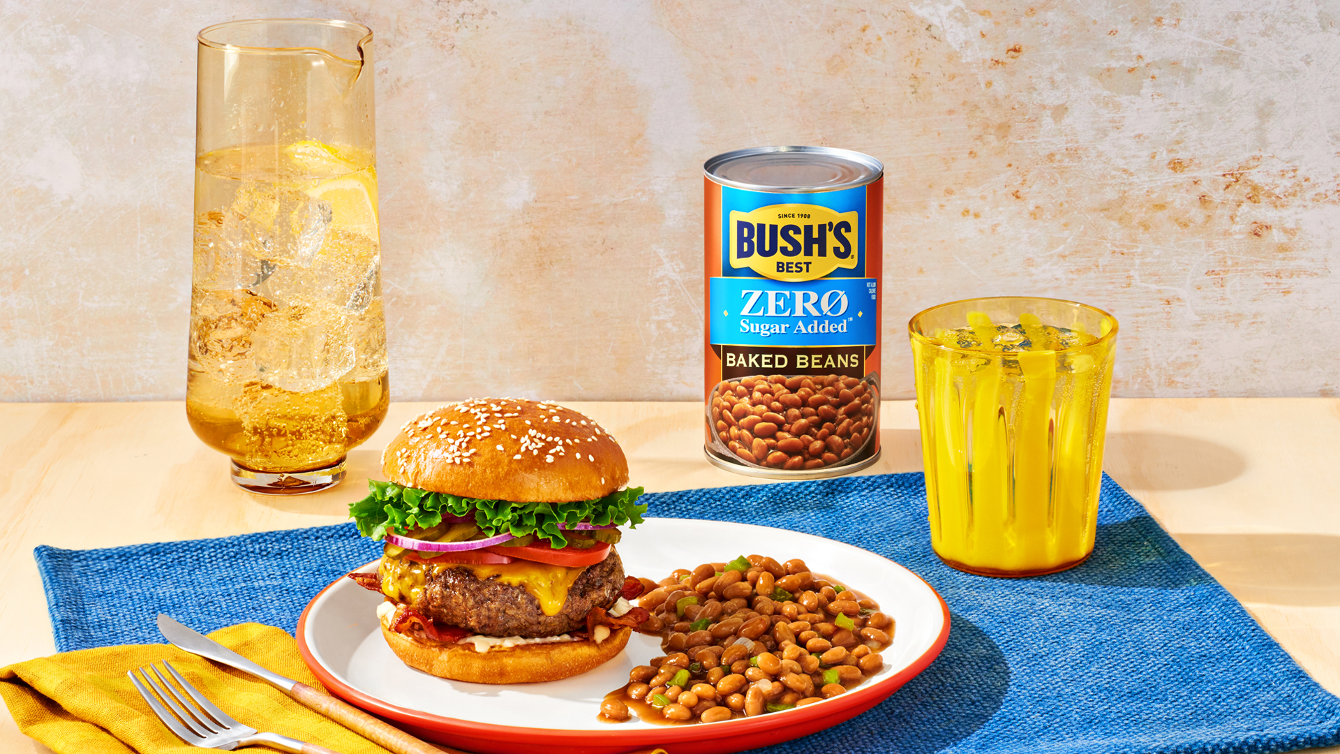 A plate with a burger and baked beans with a can of Bush's Zero Sugar Added Baked Beans on a picnic table.