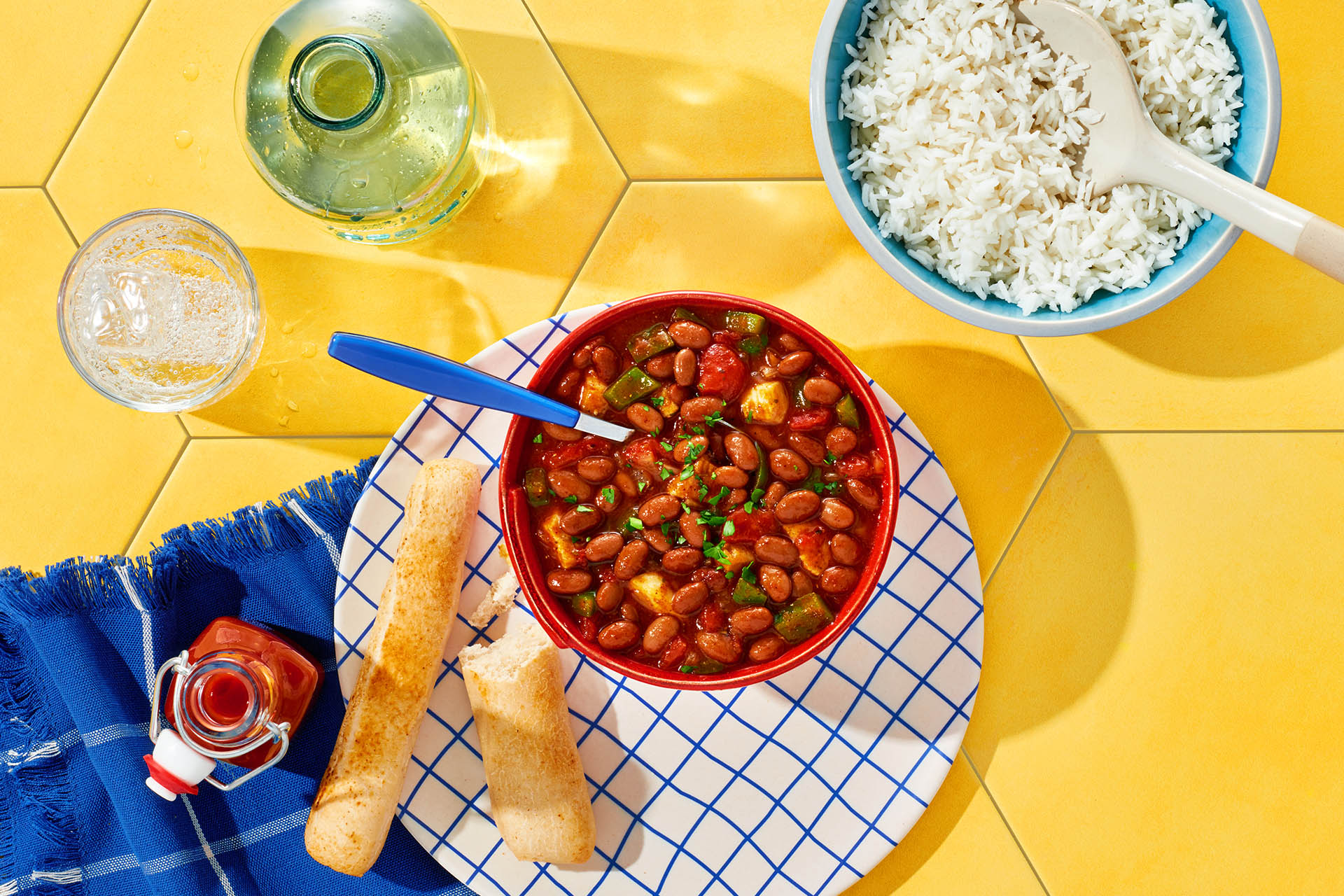 Red bowl of Hot Pepper Chicken Chili atop a yellow tile table with a side of rice and breadsticks.