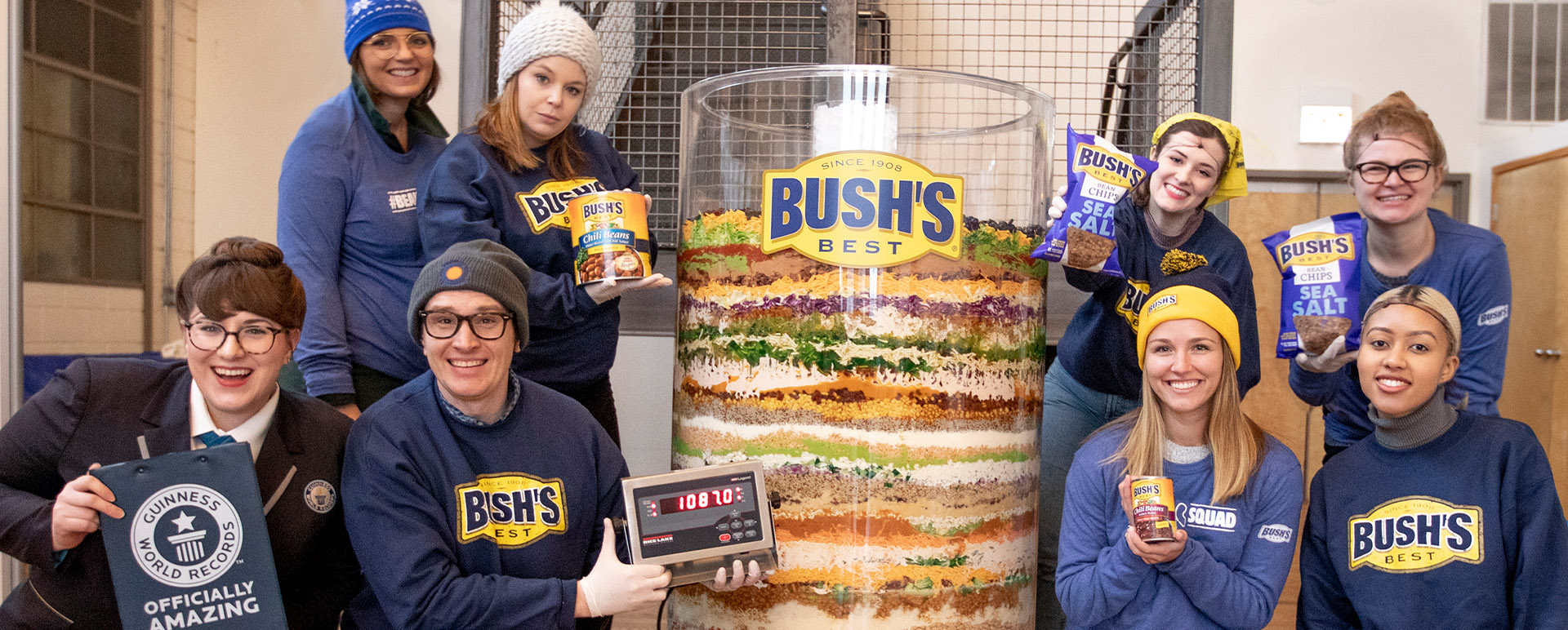 8 people posed around a large cylinder of 70 layer bean dip