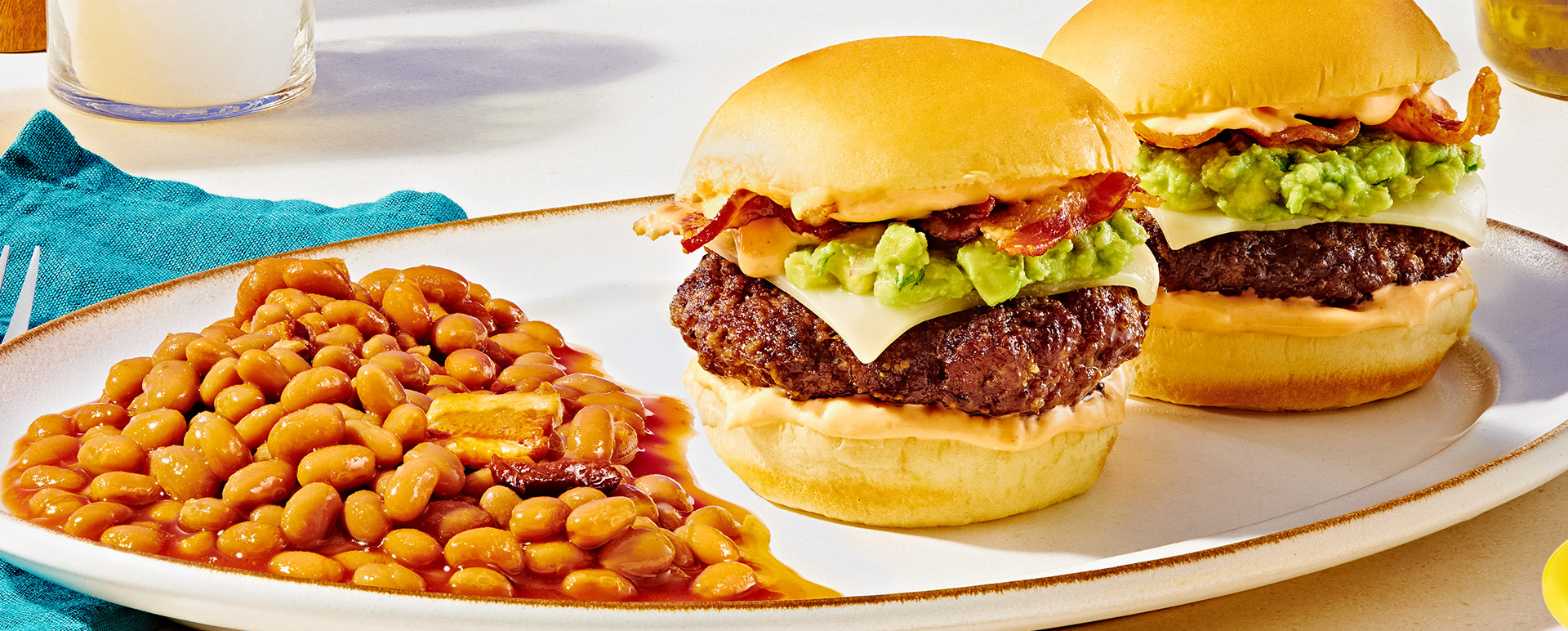 Two hamburgers topped with guacamole and mozerella with a side of baked beans with bacon on white platter